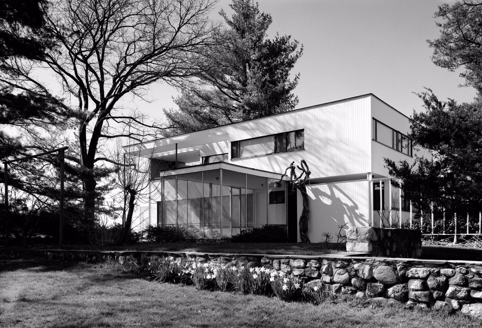 The Gropius House in Lincoln, Massachusetts: a white flat-roofed modernist house with a glass-block entry, stone wall and pines (1988 HABS photograph)