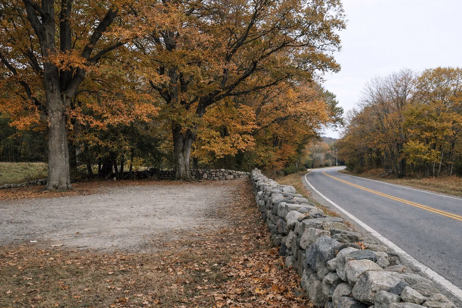 A quiet country road lined by a fieldstone wall and oaks in fall color, a small gravel parking area
